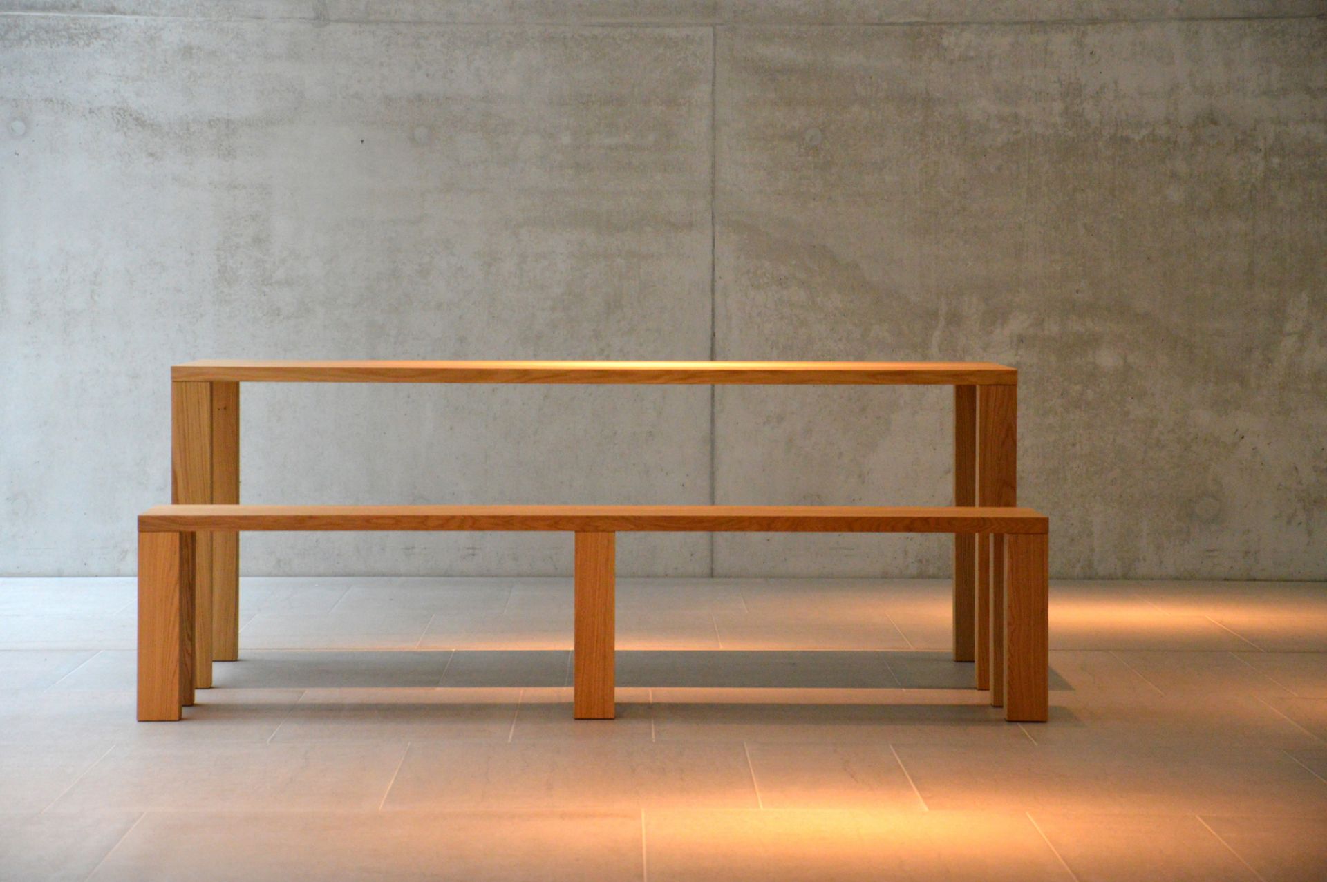 Leos Esstisch von Jan Kurtz aus hellem Holz mit passender Sitzbank vor Betonwand.